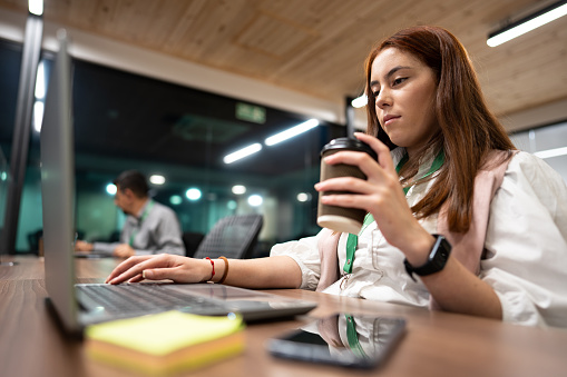 Young woman working using laptop while holding coffee cup in the office